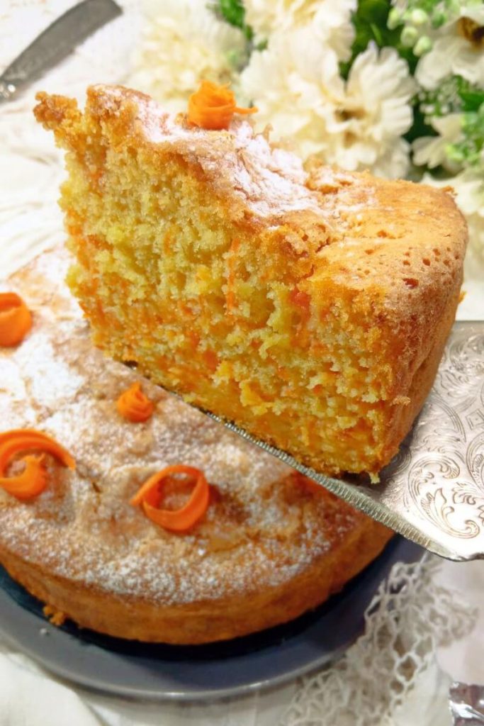Torta di carote soffice senza burro e zucchero, perfetta per colazione o dieta light Torta di carote senza burro soffice Ricette SOS – fetta sollevata con decorazioni arancioni e fiori bianchi sullo sfondo